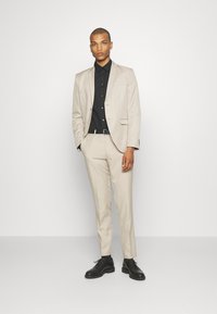 Completo beige chiaro con vestibilità su misura, blazer a un bottone e pantaloni con anteriori piatti. Camicia nera sotto, abbinata a scarpe nere.
