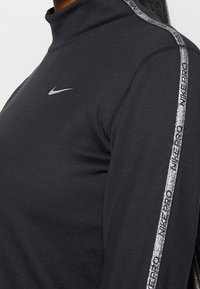 Czarny top z długim rękawem wykonany z elastycznego materiału, z półgolfem, srebrnym logo Nike oraz błyszczącym srebrnym paskiem z tekstem po boku.