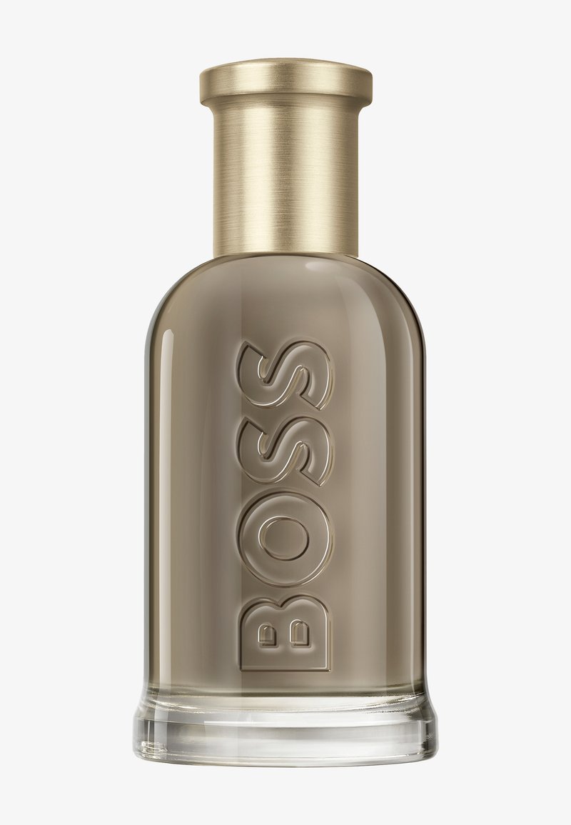 Botella de vidrio transparente de perfume Boss con líquido color topo y tapa metálica dorada, con la inscripción "BOSS" en relieve en la parte frontal.