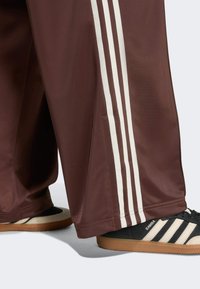 Pantalones de chándal marrón oscuro con líneas blancas a los lados, combinados con zapatillas Adidas Samba negras y beige con suelas texturizadas.