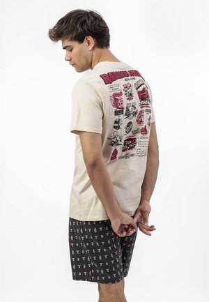 Joven de cabello oscuro que lleva una camiseta gráfica beige y pantalones cortos negros con estampado, de pie con las manos entrelazadas detrás de la espalda, mirando hacia la izquierda.