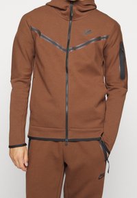 Brauner Nike Zip-Hoodie aus strukturiertem Material, mit taillierter Passform, kontrastierenden schwarzen Akzenten und einer Reißverschlusstasche am Ärmel.