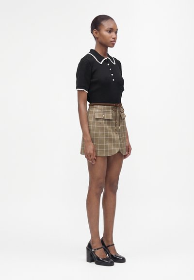 Femme avec les cheveux attachés portant un haut noir à manches courtes avec une bordure blanche et une mini-jupe beige à carreaux avec poches avant et boutons.