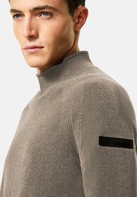 Giovane uomo con la pelle chiara e gli occhi azzurri, che indossa un maglione a collo alto color taupe con una toppa nera sul braccio superiore, mentre guarda di lato.