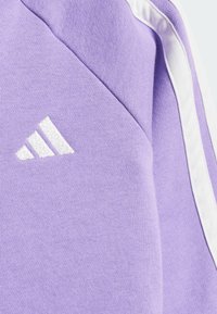 Lila pulóver textúrált kötött anyagból, fehér oldalsávokkal és hímzett fehér adidas logóval a bal felső részen.