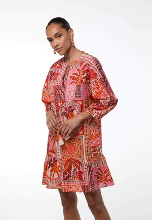Mujer con vestido holgado hasta la rodilla con estampado rosa y naranja con motivos tropicales, mangas largas y cordón con flecos en el escote.