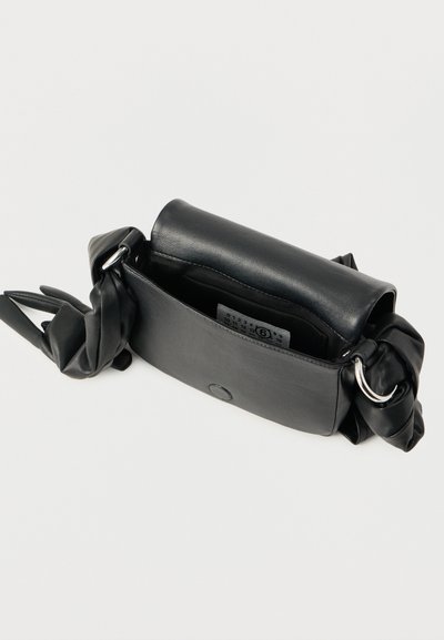 Μικρό μαύρο δερμάτινο τσαντάκι με μαγνητικό κλείσιμο, ρυθμιζόμενο λουράκι με μεταλλικούς κρίκους και ορατή εσωτερική ετικέτα σε λευκό φόντο.