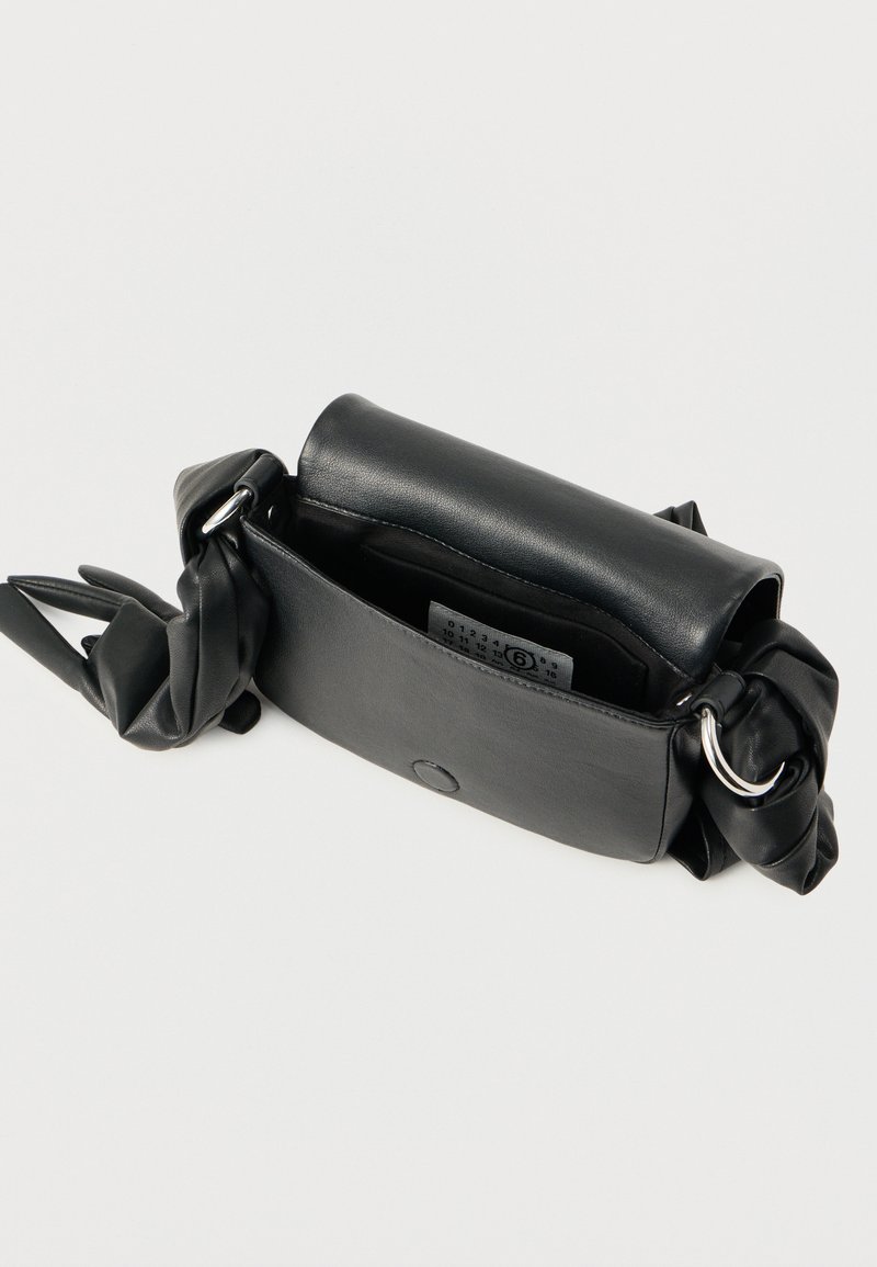 Petit sac à main en cuir noir avec une fermeture magnétique, une lanière froncée avec des anneaux métalliques, et une étiquette intérieure visible sur fond blanc.