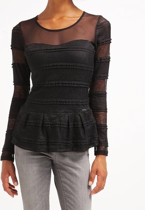 Femme portant un haut noir en dentelle à manches longues avec un col transparent et une taille peplum, assorti à un pantalon gris clair.