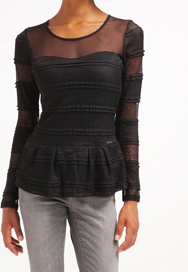 Femme portant un haut noir en dentelle à manches longues avec un col transparent et une taille peplum, assorti à un pantalon gris clair.