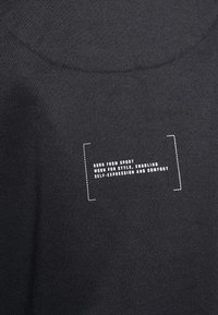 Svart sportströja med en platt textur som har vit text i en rektangulär kontur: "FÖDD UR SPORT, BÄRS FÖR STIL."