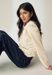 Lys cremefarvet bluse med flæse-detaljer og blondertrim, parret med mørkeblå denimjeans med synlige stikninger. Model sidder mod en neutral baggrund.