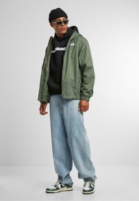 Groene lichtgewicht jas met rits, over een zwarte hoodie. Lichtblauwe losse spijkerbroek en groen-witte sneakers. Zwarte beanie en zonnebril.
