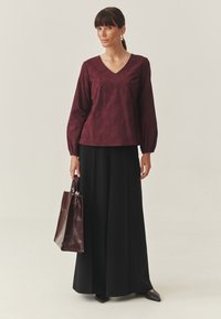 Burgundy pitkähihaiseen paita V-kaula-aukolla, jossa on teksturoitu muotoilu; yhdistetty mustaan maksihameeseen ja tummanruskeaan nahkatote-laukkuun.