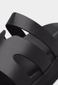 Κοντινό πλάνο ενός μαύρου σανδαλιού με φαρδιές λωρίδες και λουρί στο πίσω μέρος της φτέρνας, με ματ, ομαλή επιφάνεια.