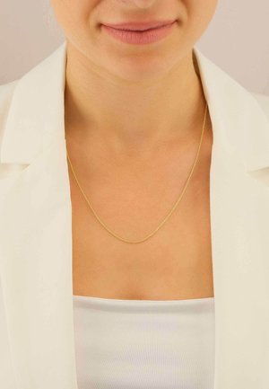 Collier chaîne en or reposant sur la peau nue, présentant un design de maillons délicats et une texture lisse, associé à un chemisier blanc et un blazer.