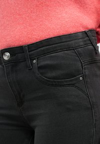 Jeans de mezclilla negra con detalles de bolsillo cosidos y herrajes plateados. Diseño de cintura alta con una textura suave. Suéter de color óxido parcialmente visible.