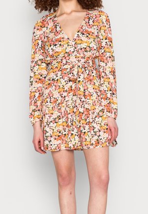 Kobieta w długim rękawie, sukience kopertowej z dekoltem w serek, ozdobionej kwiatowym wzorem w kolorach różowym, pomarańczowym, żółtym i białym, zawiązanej w talii, stojąca na jednolitym tle.