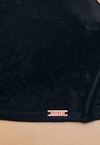 Sort blonde bh med en glat finish, intrikate blomster mønstre og en feminin guldtonet hardware-etiket med påskriften "NOIR" på bæltet.
