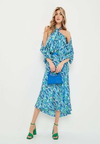 Vestido floral en azul y verde con diseño de hombros al aire, mangas fluidas, escote con lazo, combinado con un bolso azul y tacones verdes.