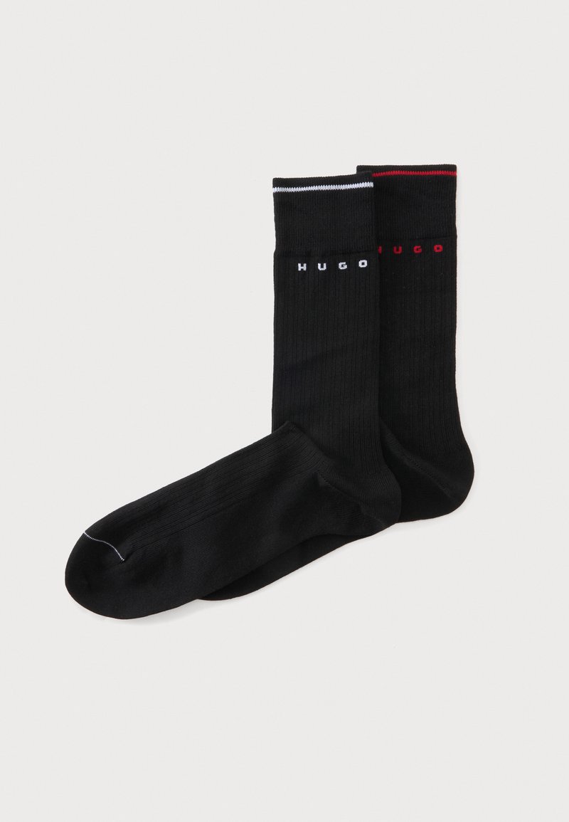 Paire de chaussettes noires montantes à texture côtelée, chacune avec le texte « HUGO » près du bord en blanc et rouge, avec des bandes assorties en haut.