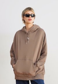 Felpa oversize marrone in tessuto morbido, con tasca a marsupio, lacci e un logo accentato sul petto. Indossata con occhiali da sole scuri.