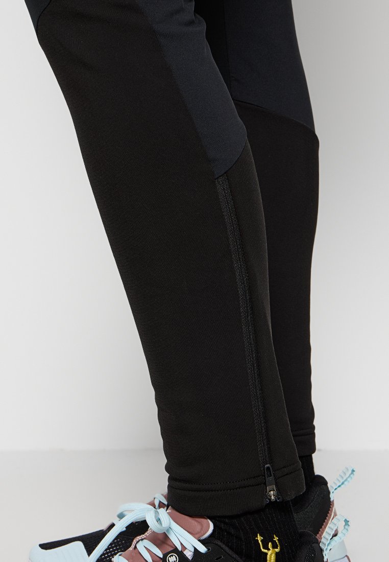 Schwarze Sporthosen mit schlanker Passform und Reißverschluss am Knöchel. Getragen mit schwarzen und rosa Sneakers mit hellblauen Schnürsenkeln und einem gelben Akzent.