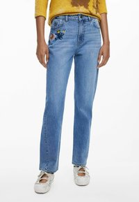 Helle blaue Denim-Jeans mit geraden Beinen und floralen Stickereien auf der Vorderseite der Tasche. Getragen mit hellen, strukturierten Schuhen.