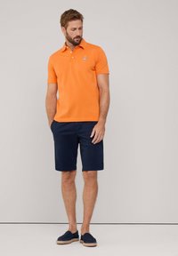 Polo arancione con logo blu, pantaloni corti blu navy a taglio dritto e scarpe slip-on blu navy con dettagli intrecciati. Sfondo neutro.