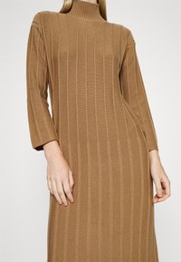 Femme portant une robe en maille côtelée camel à manches longues avec un col montant, montrée des épaules jusqu'à mi-cuisse.