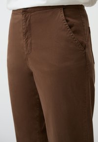 Pantaloni in cotone marrone con una texture liscia, tasche sui lati e una vestibilità rilassata, che evidenziano delicati dettagli di cucitura.