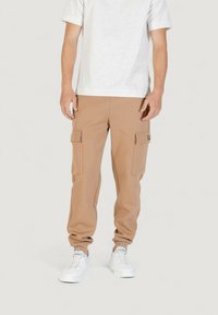 Pantaloni cargo beige realizzati in tessuto morbido, con due tasche frontali, orli a costine e una vestibilità ampia, abbinati a sneakers bianche e una maglietta grigia chiara.