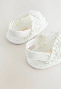 Bílé látkové sandály s krajkovými detaily, nastavitelnými popruhy a měkkou podrážkou. Navrženo pro kojence, s kulatou špičkou.