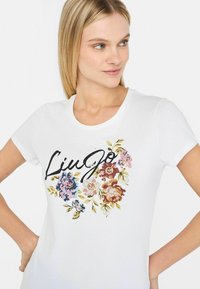 Hvid t-shirt med kortærmer, der har et blomstermotiv i pink, blå og guld, med "Liu Jo" tekst i sort henover designet. Blødt bomuldsstof.
