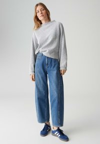 Donna che indossa un maglione lavorato a maglia grigio chiaro, jeans a gamba larga a vita alta di colore blu e scarpe da ginnastica blu con strisce bianche, in piedi su sfondo neutro.