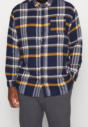 Mand iført marineblå, gul og hvid ternet skjorte med knapper og forlommer samt grå bukser, stående med afslappede hænder.