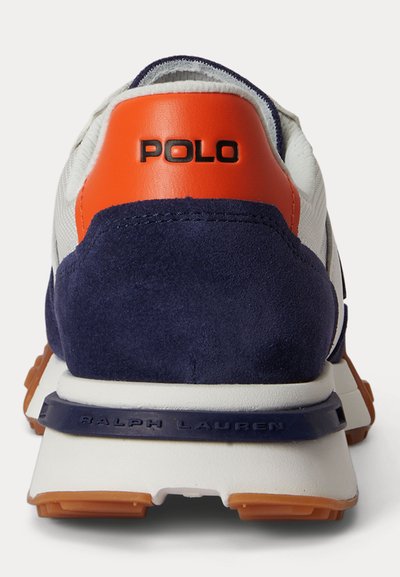 Πίσω μέρος λευκού αθλητικού παπουτσιού με ναυτικό σουέντ, πορτοκαλί ταμπελάκι στη φτέρνα με την επιγραφή "POLO" και ναυτική διακοσμητική λωρίδα στη μέση της σόλας με ανάγλυφη την επιγραφή "RALPH LAUREN".