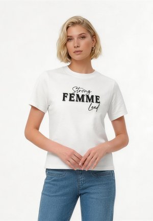 Blondynka w białej koszulce z napisem "Strong FEMME Lead" i niebieskich dżinsach, stojąca z rękami luźno złączonymi przed sobą.