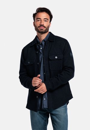 Homme avec une barbe portant une veste boutonnée bleu foncé sur une chemise bleu marine et un jean bleu, se tenant devant un fond blanc uni.