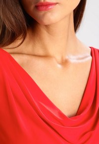 Czerwony top z karczkiem, o gładkiej teksturze, drapowanej tkaninie i delikatnym połysku. Zbliżenie na dekolt i ramiona, ukazujące kontury i subtelny blask.