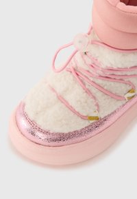 Bota de invierno rosa con parte superior blanca y esponjosa, detalles metálicos en rosa y un sistema de cordones elásticos. Cuenta con un puño acolchado para mayor calidez.