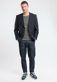 Blazer bleu marine porté sur une chemise à rayures noires et blanches, associé à un pantalon en denim foncé et des baskets bleues avec des accents blancs. Design simple, look décontracté.