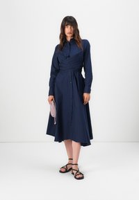 Mörkblå skjortklänning med långa ärmar, knappfront, insvängd design vid midjan och flödande kjol. Bärs med svarta sandaler och en liten rosa väska.