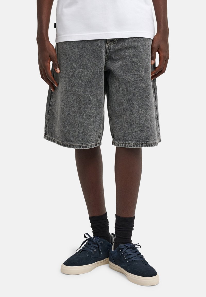 Short en jean gris foncé à coupe ample, ourlet effiloché, et design standard à cinq poches, associé à des baskets foncées et des chaussettes noires.