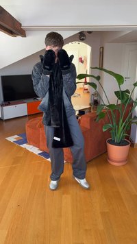 Mustast karvast sall käte ümber, sobivate mustade kinnastega. Hall teksajakk ja hallid püksid, seisab elutoas, kus on taimi ja mööblit.