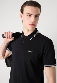 Čierna polo košeľa s bielym logom "BOSS". Má klasický golier so pruhovanými akcentmi, krátke rukávy a hladkú textúru látky.