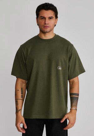 Mand med mørkt hår iført olivengrøn oversized T-shirt og sorte bukser, der viser tatoveringer på begge underarme, står mod en ensfarvet baggrund.