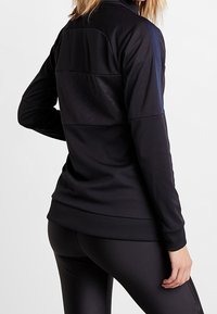 Femme portant une veste de sport noire à manches longues ajustée et un legging noir, se tenant de dos face à l'appareil photo contre un fond uni.