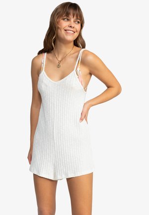 Jonge vrouw die glimlacht, gekleed in een witte geribbelde mouwloze romper met zichtbare bloemenbandjes eronder en een ronde hangerketting.