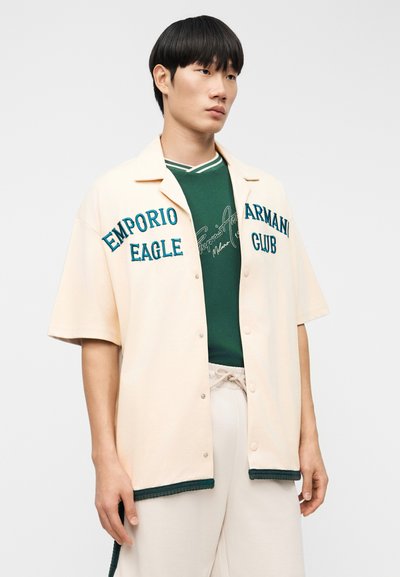 Μπεζ πουκάμισο με κοντά μανίκια και κουμπιά, με πράσινα κεντημένα γράμματα, φορεμένο πάνω από ένα σκούρο πράσινο πουκάμισο με λευκές λεπτομέρειες, συνδυασμένο με ανοιχτόχρωμο παντελόνι.